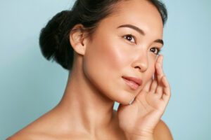 Skin care. Woman with beauty face and healthy facial skin portrait. Beautiful asian girl model with natural makeup touching glowing hydrated skin on blue background closeup. High quality image Revive Laser and Skin Clinic